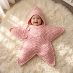 Smiling baby wrapped in a blush pink crochet star baby wrap with the hood up, lying on a cream rug